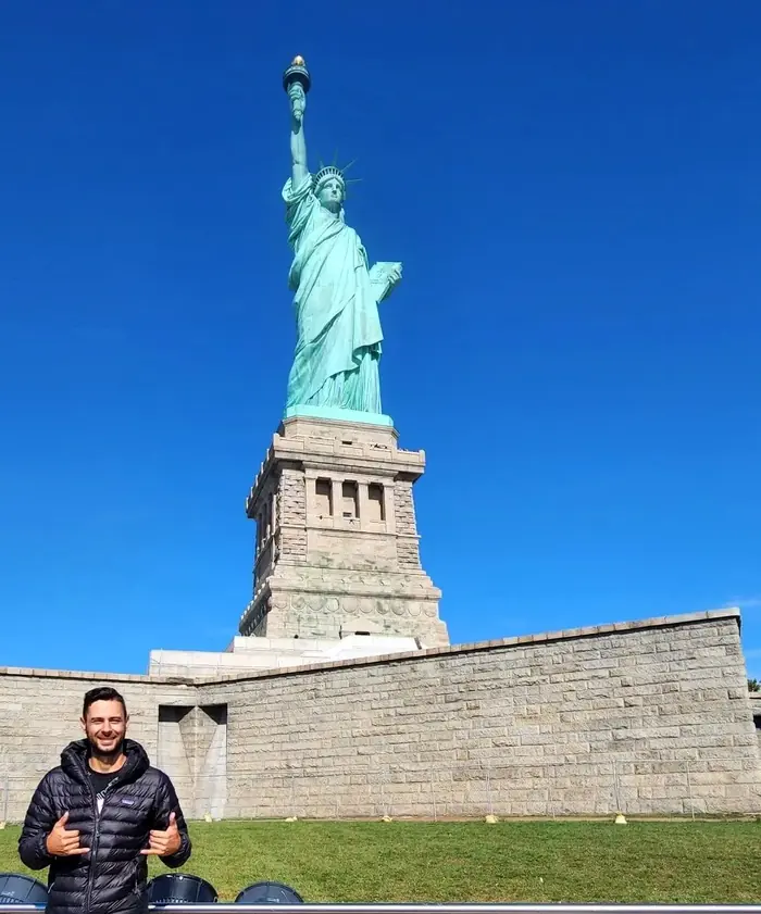 Kristiyan Rukov smiling in a puffer jacket in front of the Statue of Liberty in New York